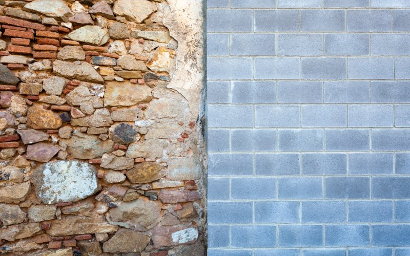 Photo of a wall divided in two. The left side is chaotically made of rocks and thin red bricks. The right is orderly grey concrete bricks.