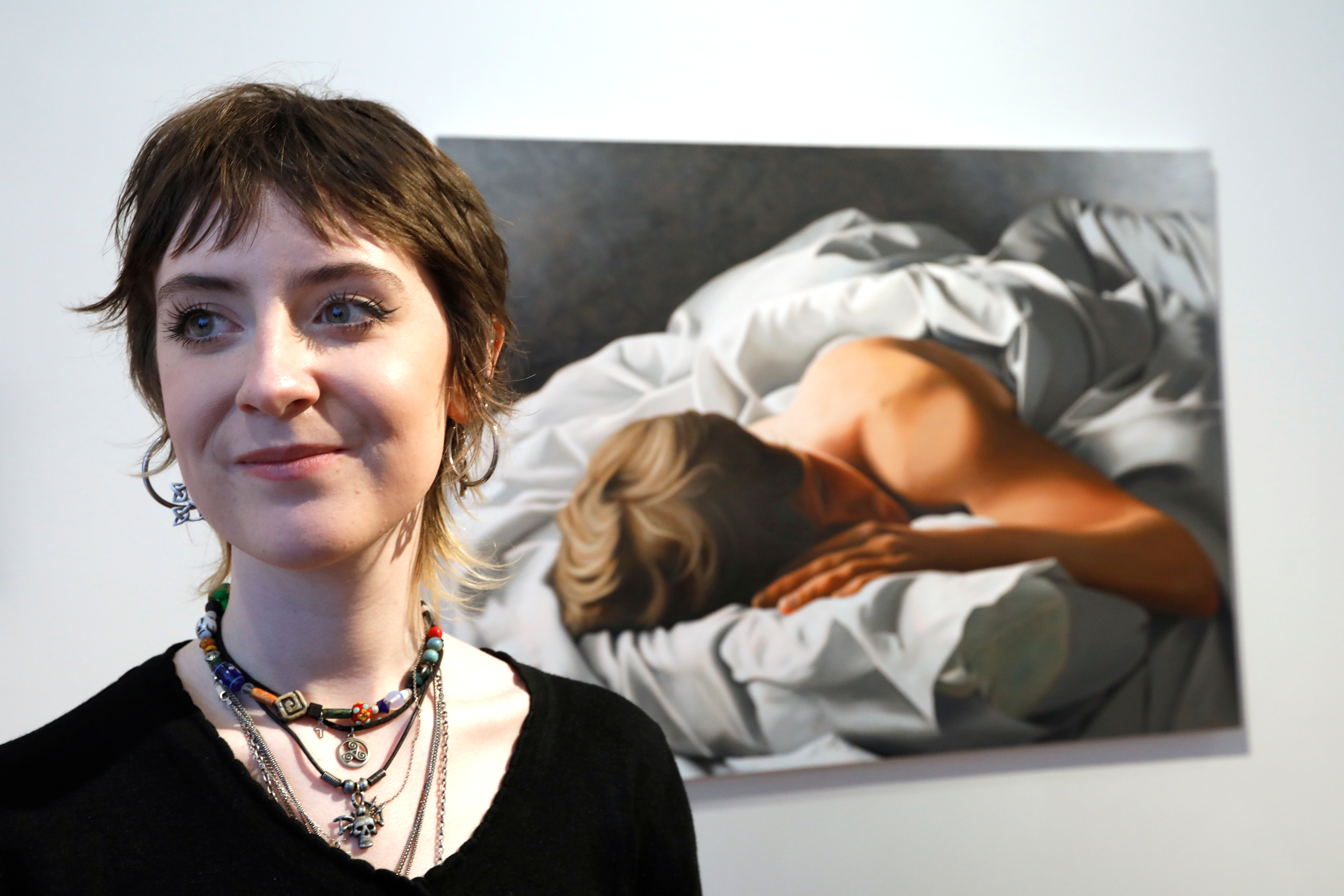 The artist, Eimear, is photographed near the camera and behind her is her painting of a man asleep in a tussled white linen duvet and bed. Eimear has short brown hair and is looking to the left.