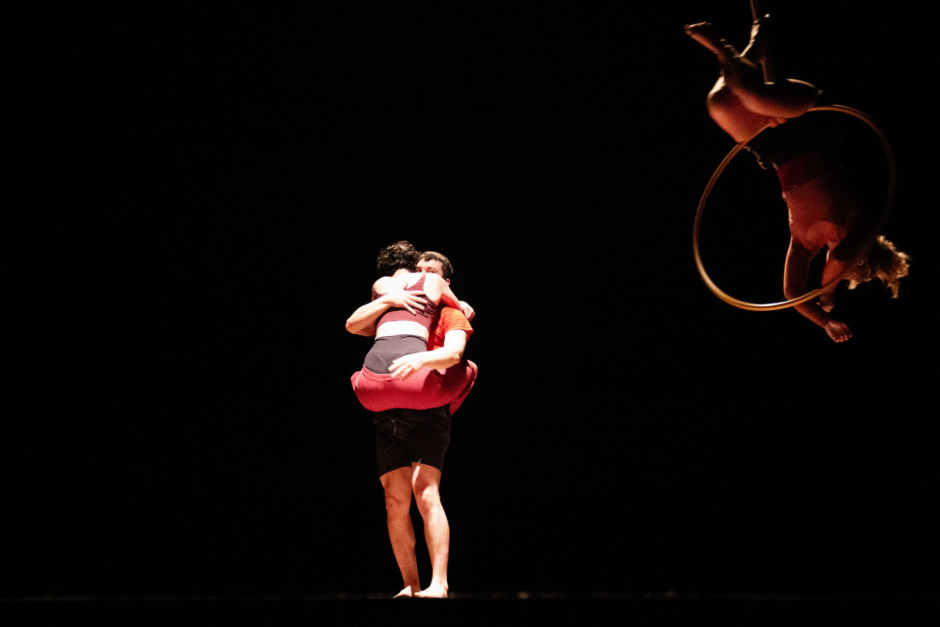performance photo of Janas. A man and woman wearing exercise shorts and tops. The woman is held by the man in a hug with her legs wrapped around his waist. They are brightly lit against a black background.