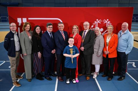 A group standing on an indoor athletics track with a red Special Olympics backdrop and a young person holding a torch at the front.