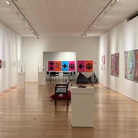 A long room with bright coloured art on either wall. In the middle of the room are plinths that hold small sculptures.