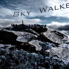 a rocky outcrop with scrub and grasses is in the foreground of this photograph. a number of people may be seen standing on the top of the rock in the distance, framed against a dramatic sky with clouds. The words Sky Walkers float in the clouds. The entire image is filtered with a blue tinged infra red filter. 