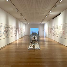 Photo of the Gallery in dlr LexIcon. A long white room with paper artworks pinned to the walls on the left and right of the room. In the middle of the room is a long metal cage containing lots of small items.