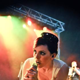 Julie, a singer with drak hair tied up, is holding a microphone and leaning down into it. She is shown on a stage with yellow, red and green lights behind her. These are attached to a lighting rig at the top of the frame.