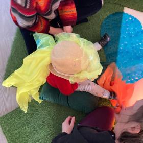 A colourful picture of a small child sitting on the floor with two adults sitting either side. the child has a yellow piece of fabric over their head.