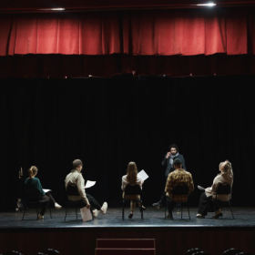 6 people are on a dark stage with a red curtain above. 5 men and women are sitting with their backs to the camera. infront of them is a man standing. some have paper in their hands that looks like scripts.