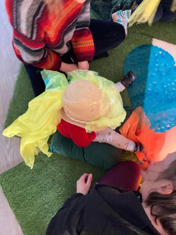 A colourful picture of a small child sitting on the floor with two adults sitting either side. the child has a yellow piece of fabric over their head.