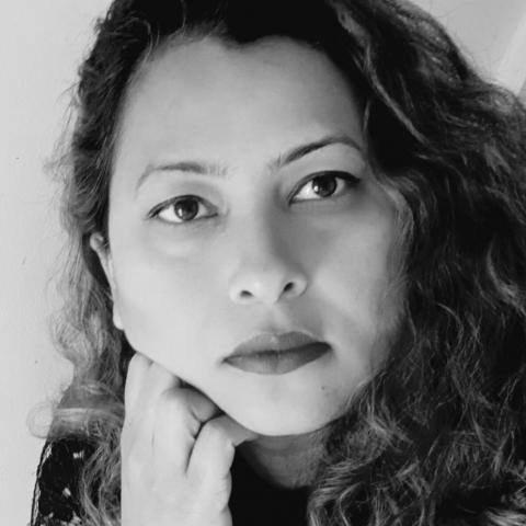 Hina Khan a black and white photo portrait of the artist Hina Khan. Hina is looking at the viewer, her chin is resting on her hand. She has long curly brown hair.