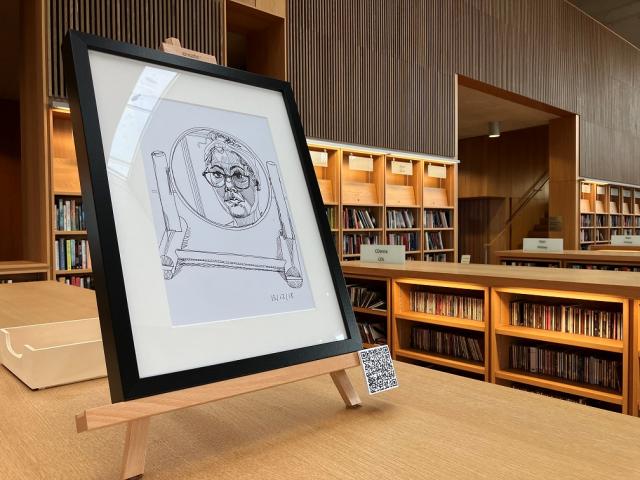 Self portrait drawing in a round mirror. drawing is in a black frame on an easel on top of a library bookshelf