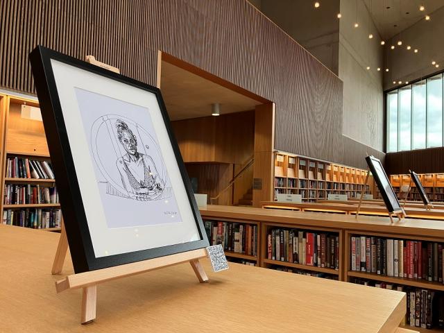 Self portrait drawing in a round mirror. drawing is in a black frame on an easel on top of a library bookshelf