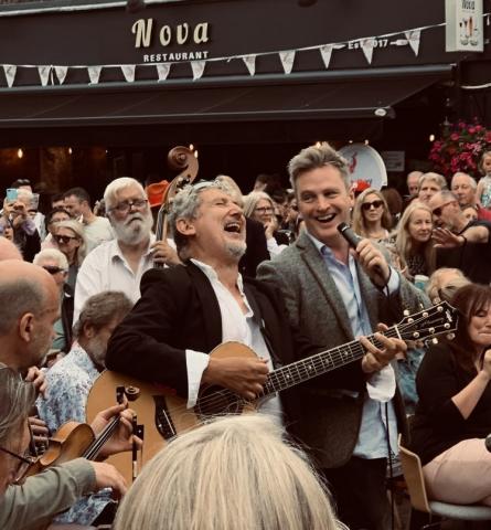 Drazen Derek and Simon Morgan performing in the middle of a large crowd of people. Drazen, on the left is playing guitar, and Simon is holding a microphone