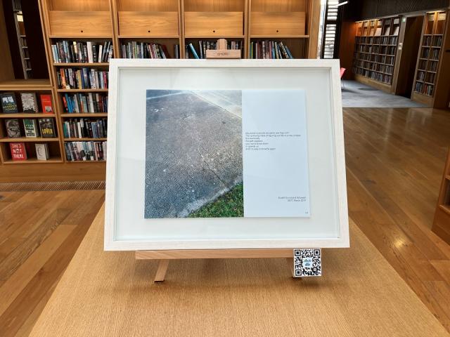 A framed picture is on top of a library bookcase. It contains two images. One is a concrete pavement beside a small patch of grass and the other is a quote from Ellie Berry's journals.