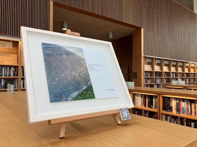 A framed picture is on top of a library bookcase. It contains two images. One is a concrete pavement beside a small patch of grass and the other is a quote from Ellie Berry's journals.