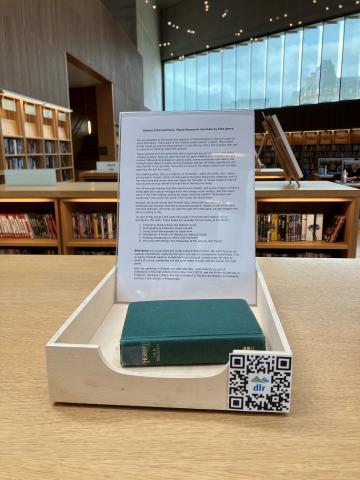 A wooden box containing a book on top of the library bookshelf. At the back of the box is a perspex stand with information about the artworks.