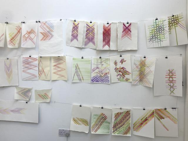 three rows of small painting are hung across a white wall from left to right. They are clipped onto a string with small bullclips. The paintings are abstract with geometric shapes and lines in various colours, mainly greens, purples, yellows and reds