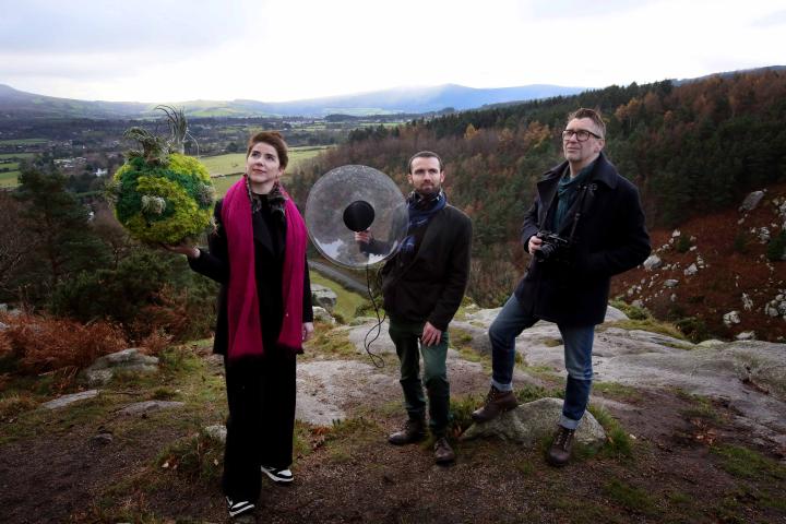 Meadhbh O'Connor wearing black clothes and bright pink scarf holds a moss covered circular object in her right had; Robert Coleman wearing dark clothes holds a microcphone which is encased in a clear plastic circle; Martin Healy in dark clothes, holds a camera. They stand facing the photographer, standing on a rocky surface with mountains behind them in the distance