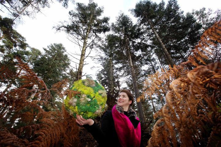 Méadhbh O'Connor, wearing black with a brigh pink scarf, is standing in the foreground of the photograph facing the camera. the camera angle is low and there are tall pine trees behind her. She holds a bright green moss covered ball in her right hand