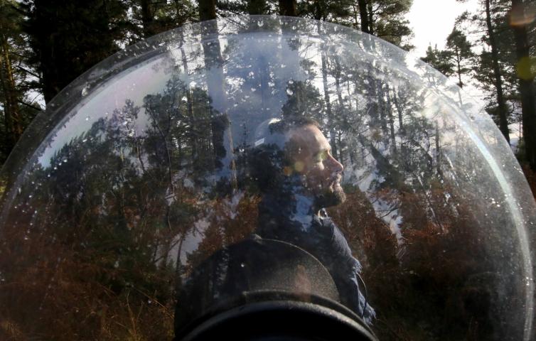 Robert Coleman stands with his side profile to the camera, looking into the distance. he is wearing headphones and there are trees behind him. The photographer has taken the image looking through a plastic circle and this gives the photograph some blurring and distortion