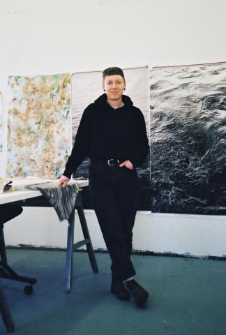The Artist Roisin White is standing in an artist studio dressed in black with her legs crossed. Behind her are large photo prints of the sea.