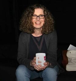 Maura Campbell sits centre frame facing the camera. she is in a relaxed pose with a coffee cup held in both hands. She wears glasses with dark frames and has curly brown hair to her shoulders, she is smiling