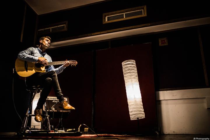 Chris sits on a high stool to the left of the frame. He is playing an acoustic guitar with his eyes closed. To the right of the frame there is a lamp and the photo is shot from a low angle looking up at Chris