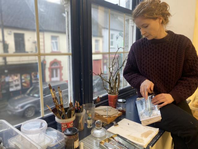 Joanna, to the right of the frame, has red/brown hair tied up. she wears a dark cable knit jumper and is looking down at a sketchpad resting on the windowsill. she sits on a wide windowsill painted  a dark grey/black and has a selection of brushes, containers and small branches on it. in the distance through the windown behind her, is a street and a row of houses and shops