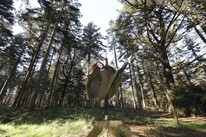 Laura, a dancer with long dark blonde hair, stands on a tree stump on one leg. her two arms are reaching overhead and her other leg is held high. she wears an olive green dress and cape. she is in the middle of a forest clearing with tall pine trees behind her