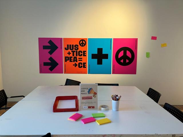 A large table with bright coloured post it pads, pencils and paper. On the wall behind the table are 4 solid colour posters that have arrows, plus and peace signs and the words Justice and Peace