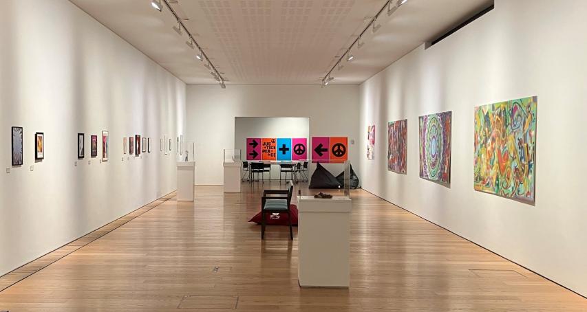 A long room with bright coloured art on either wall. In the middle of the room are plinths that hold small sculptures.