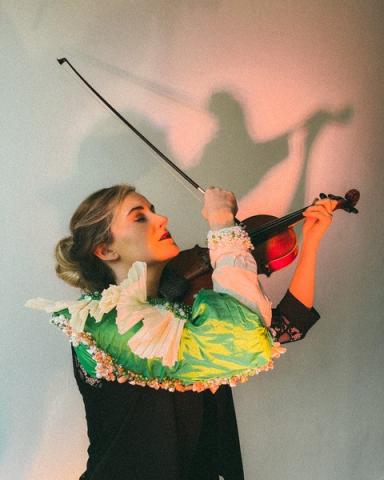 Jane wears a black top with brightly coloured sleeves of green, yellow and cream. Her blonde hair is tied back. Her eyes are closed, she holds a violin in her left hand and the bow in her right. she is pictured with her head facing upwards against a plain white background