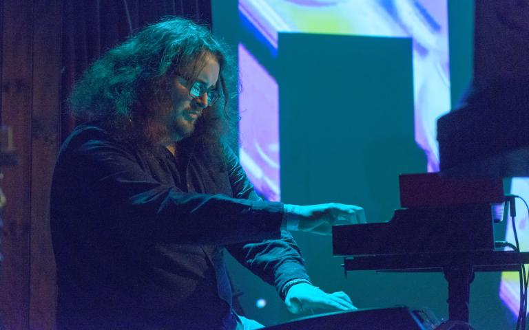 Luke is seated at a piano, he is to the left of the image. He has shoulder length dark hair and wears glasses.The image is shot from a low angle looking up at Luke and shows him from the waist up, with the piano to the right of the image. The photograph has an overall blue/green filter with some purple shot through it