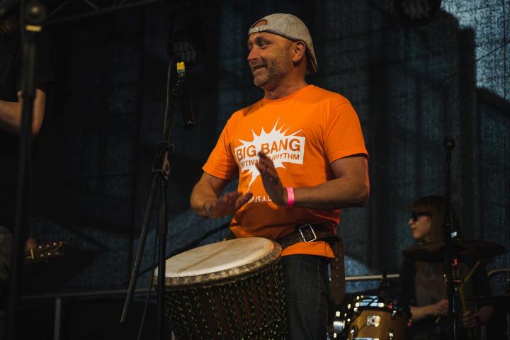 Brian, wearing a light brown cap turned backwards on his head and a bright orange t-shirt, sits centre stage. He is playing the bodhran and smiling