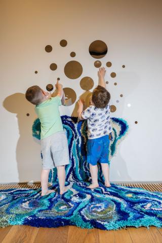 Two small boys are reaching and pointing at round mirrors stick to a while wall. they are standing on a blue swirly carpet that goes up the wall 2 feet.