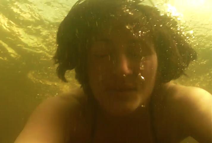 a woman's face under yellow tined water. Her eyes are closed and bubbles traipse from her mouth up to the surface of the water rippling above