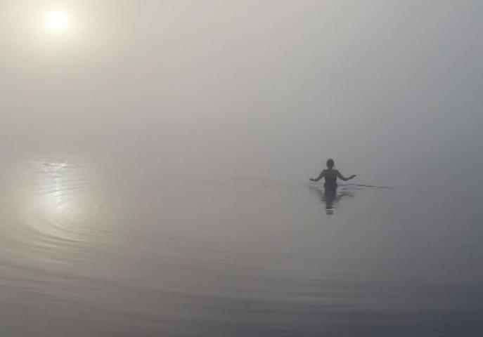 The silhouette of a woman who is wading into still water on a misty day. the sun is glowing through the mist on the left