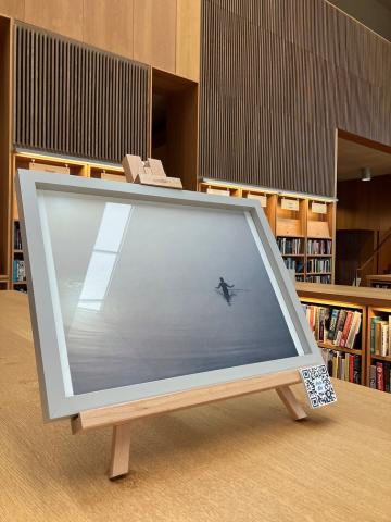 a framed photo on top of a chest height bookcase shows a misty body of water with a person wading into the water.