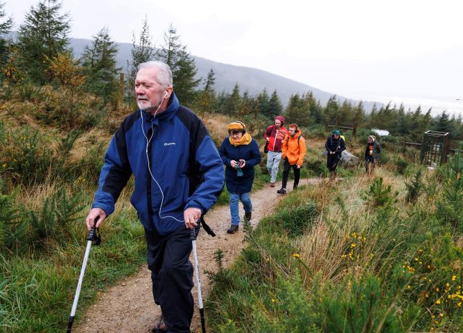A boardwalk in a forest setting. Several people are walking on the boardwalk led by a man using walking poles who is pictured in the foreground. There are trees behind the boardwalk and hills in the distance. The walkers are wearing raincoats and the light is dull and misty.