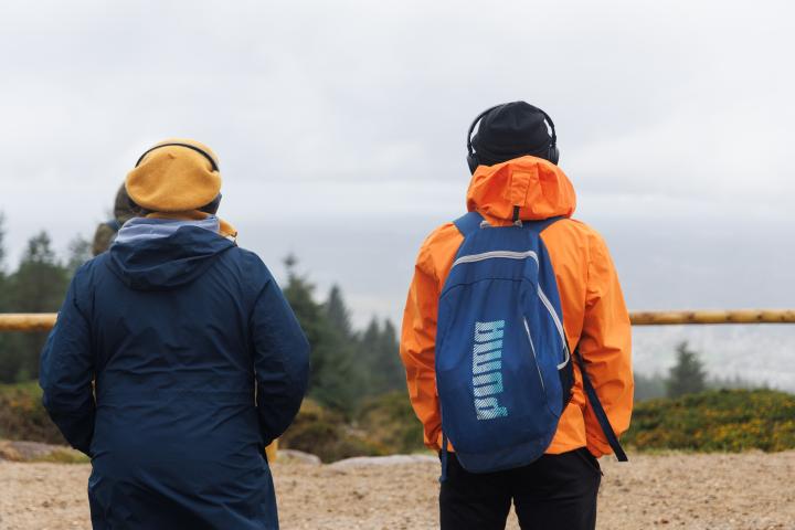 two people stand with their backs to the camera. they are looking out towards the sea from a high elevation in a forest. The lady wears a yellow hat and navy raincoat, the man wears a black hat and orange raincoat and has a backback on his back