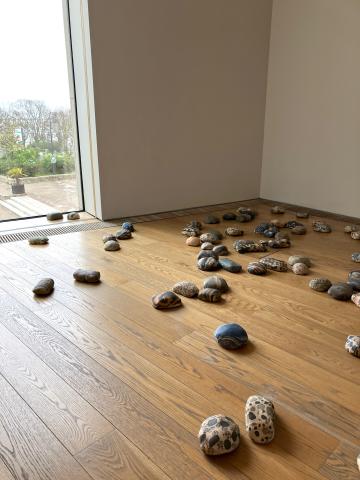Photo of what look like many smooth rocks sitting on a wooden floor. In the background is a floor to ceiling window looking down on the metals in Dun Laoghaire.
