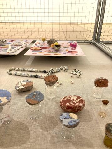 Photo of inside a cage in a gallery. There are small coloured pieces of clay displayed on top of small glass jars. There is a string of black and white beads.
