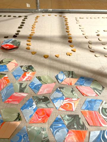 Photo of inside a cage in a gallery. Close to the camera are pieces of coloured paper cut into shapes which are placed to look like 3 dimensional cubes. In the background are small pieces of clay placed in the shape of a necklace.