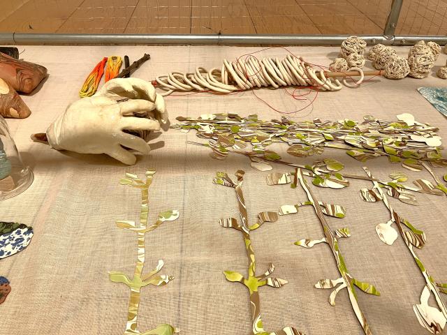 Photo of inside a cage in a gallery. Close to the camera are cut out pieces of paper in the shape of leaves on a vine. Behind them is a ceramic set of hands which look like they are holding onto something. Behind them are a row of ceramic rings.