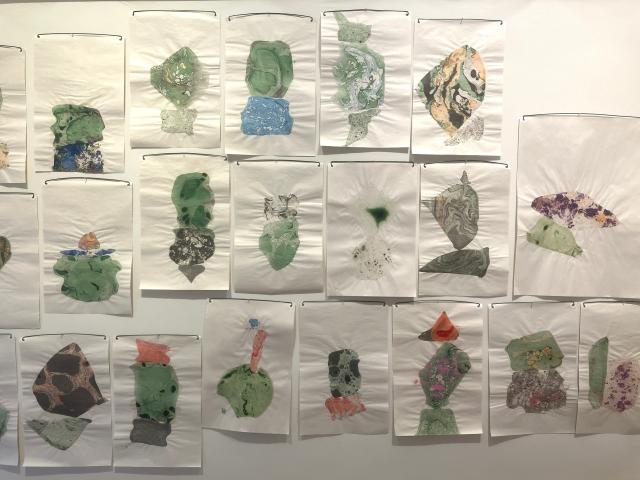 Photo of many pieces of paper pinned to a white wall. On the paper are different shapes and colours. There is a lot of green in this section.
