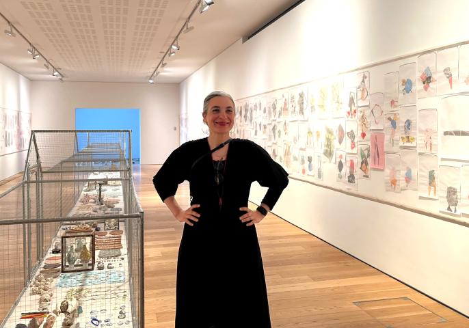 The artist Vanessa Donoso López, is wearing a black dress with her hands on her hips, facing the camera. She is standing in a gallery next to her artwork which is a long metal cage on the ground which displays small curiosities and on the wall are many pinned pieces of paper with colourfull images on them.
