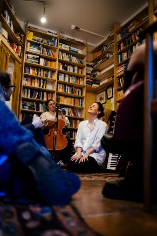 Aoife, a singer, is sitting on the floor with her eyes closed and head tipped back. She is wearing a white shirt and dark trousers and her hands are resting on her thighs. beside her another musician is playing a cello, she is also wearing a white shirt. There are high walls of bookcases behind them and in the foreground is the arm of an audience member also sitting on the floor, wearing blue