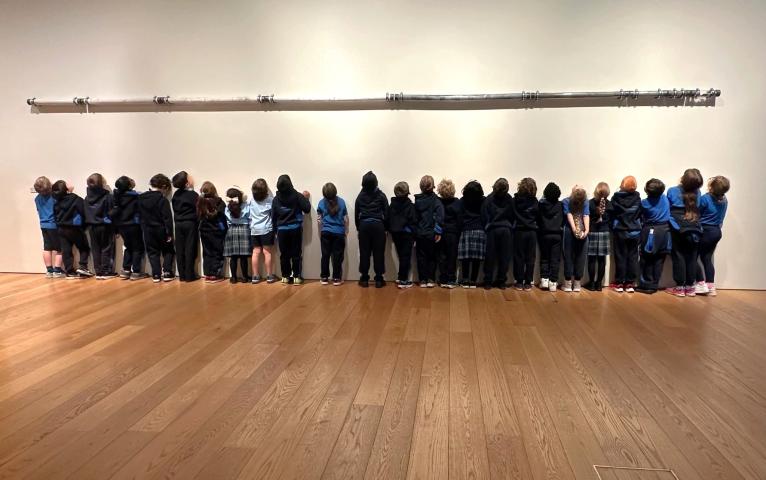 a row of young children, in school uniforms, are facing a white wall looking up at a long dark tube that is attached to the wall.