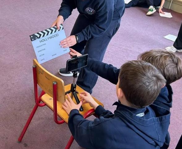 two boys are kneeling in front of a childs school chair. on the chair is a smart phone on a tripod. The phone is facing a film clapperboard being held by another boy.