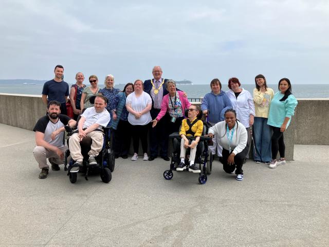 A group of people are standing along the seafront with a wall behind them. The group is made up of men and women, and two people are in wheelchairs. In the middle is a man with a large gold chain around his neck that indicates he is the Cathaoirleach of Dún Laoghaire Rathdown county.