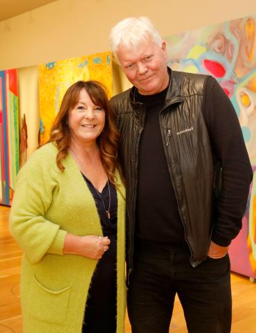 The artists Karen Ebbs is standing next to artist Dave Madigan in front of large colourful abstract paintings