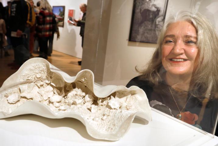 A porcelain sculpture of a large, open, shell like object. Inside the shell are many small porcelain flowers. Behind the sculpture is the smiling artists Andrea Kavanagh.